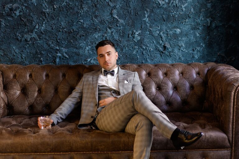 front-view-handsome-bridegroom-with-beard-wearing-trendy-suit-sitting-leather-sofa-luxury-apartment-holding-glass-filled-with-beverage-looking-camera-min